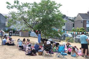 A woman standing talks under a tree with a small crowd sitting on the grass and listening